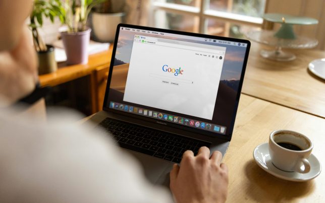 An adult using a laptop indoors, browsing Google at a wooden table with coffee.